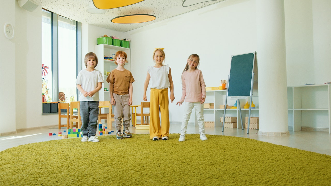 Four children are standing and smiling in a brightly lit classroom with white walls and a green round carpet. Behind them are shelves with colorful toys, a small table with chairs, and an easel. The room has large windows and ceiling decoration with orange panels.